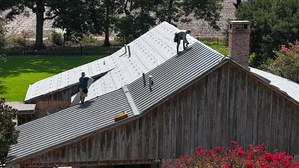 Drone shot of roofing crew installing gray metal roof panels and synthetic underlayment on a ranch-style house next to a pool.